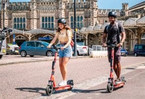Two people riding voi e-scooters in front of Temple Meads Railway Station.