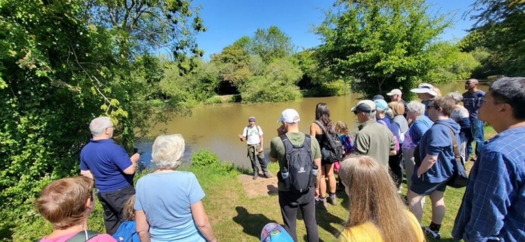 Photo of walk attendees gathered beside a lake while the walk leader talks to them about local wildlife and the environment.