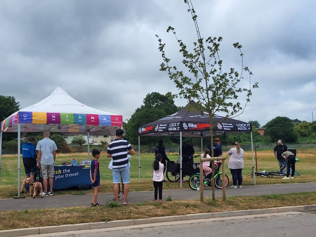 Photo of a sustainable travel roadshow set up beside the path at Beaufort Park, with the roadshow team, police bike marking, and Dr Bike offering cycle checks