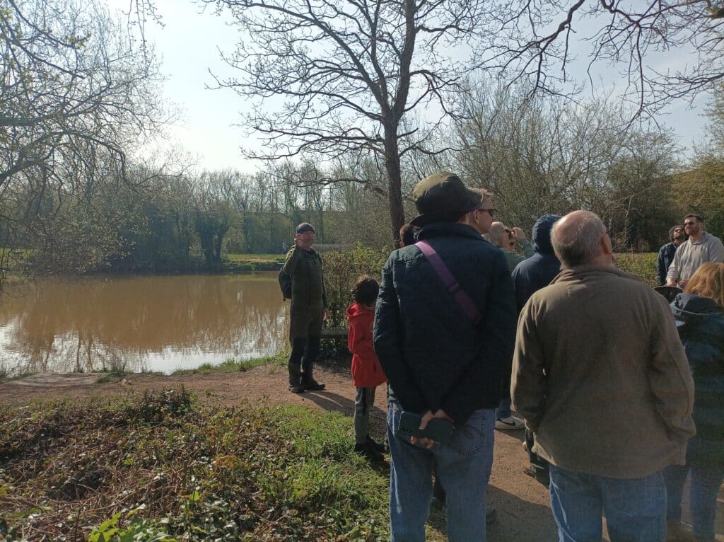 Photo of the walk leader standing beside a lake, pointing to a hole in a tree trunk used as an ideal bat habitat.