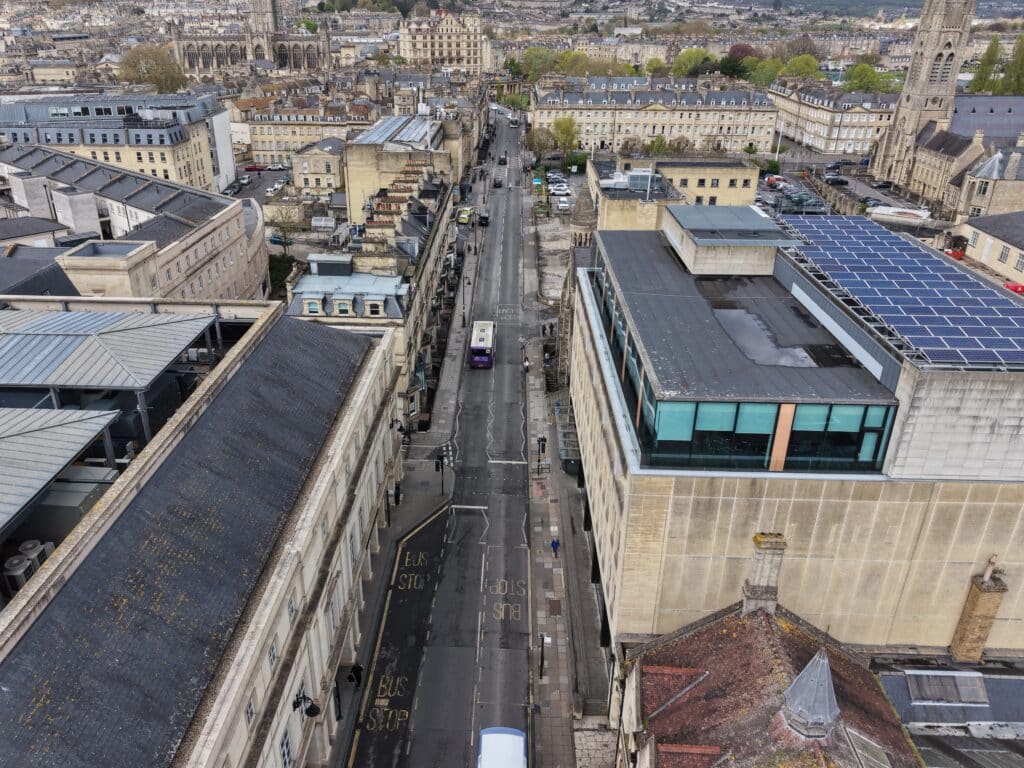 A high-angle view down Manvers Street in Bath showing a long, straight road flanked by stone-built Victorian and modern buildings; pedestrian pavements, parked cars and a single bus are visible, with the city’s skyline and church tower in the background under a pale sky.