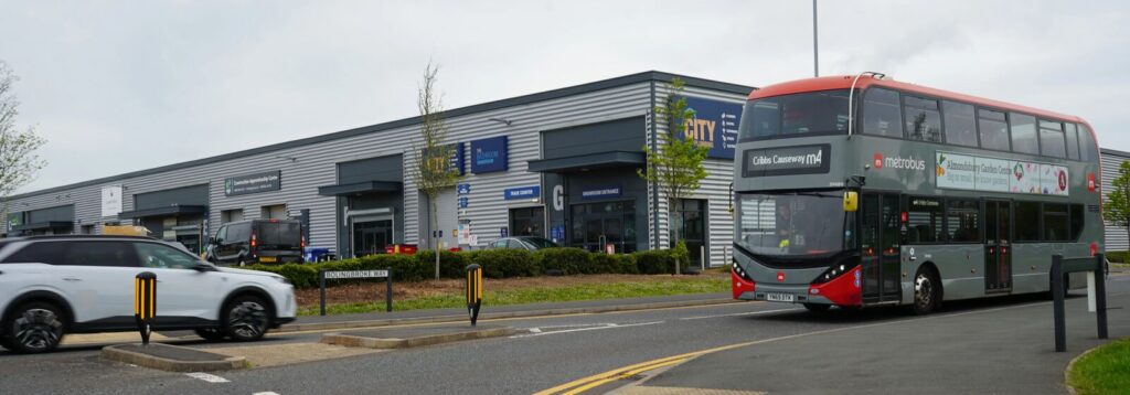 A wide, rectangular photo showing a modern business park storefronts in the background with a two-story red-and-gray double-decker bus turning on a road at right; a white SUV waits at a pedestrian island at left, low curbs and small trees line the landscaped central strip, and the sky is overcast.