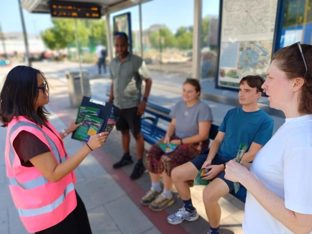 A travel advisor in a high-visibility vest speaks with a group of passengers at a bus stop, handing out travel information leaflets. A real-time departure display and local transport map are visible in the background.